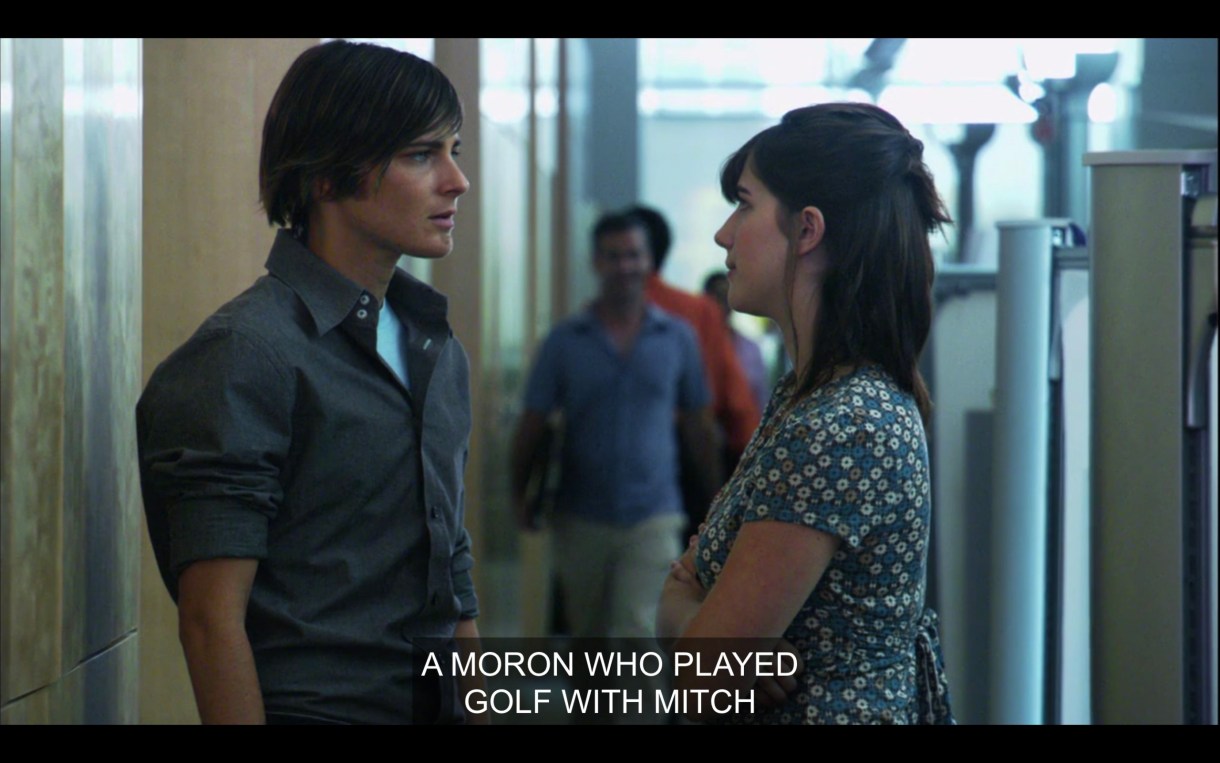 Max (wearing a gray dress shirt) is standing in his work office facing a brunette women in a blue and green dress. "A moron who played golf with Mitch."