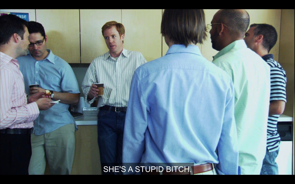 A bunch of guys (including Max, whose back is to the camera) standing around in a circle in the office break room. One of them says, "She's a stupid bitch."