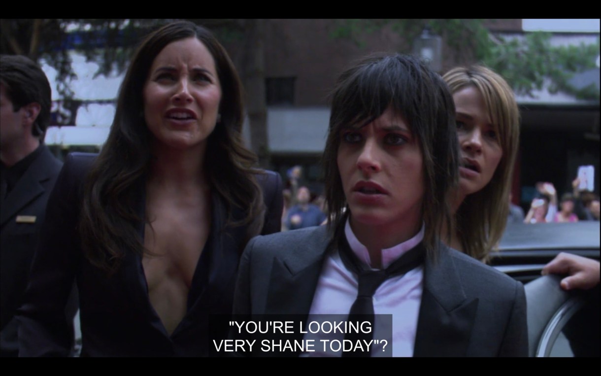 Helena (wearing a high-cleavage black jacket), Shane (wearing a skinny black tie over a white shirt and a black blazer), and Alice (peaking over Shane's shoulder) first look at Shane's billboard. Helena reads, "'You're looking very Shane today?'"