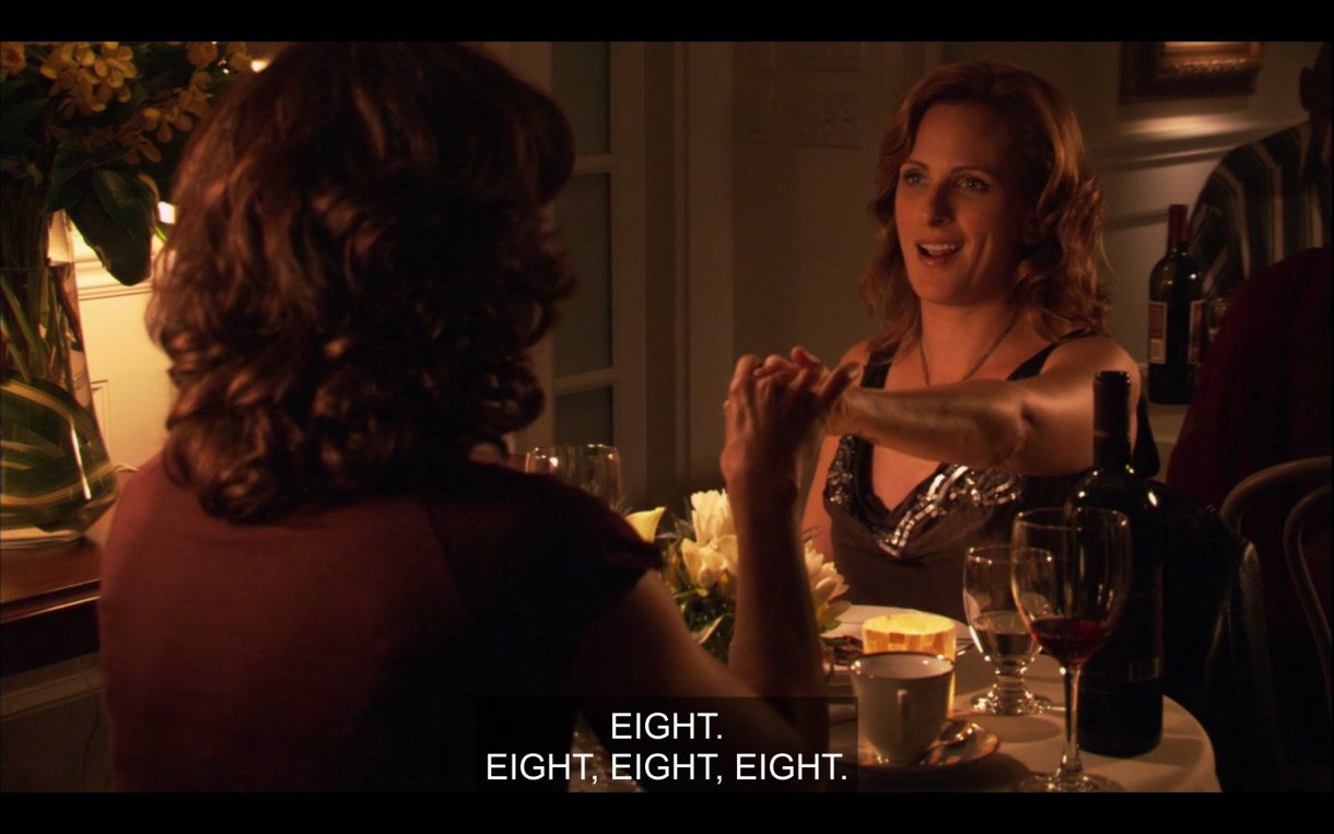 Bette (wearing a brown top, her back to the camera) sits across a dinner table from Jodi. They're holding hands across the table. "Eight. Eight, eight, eight."