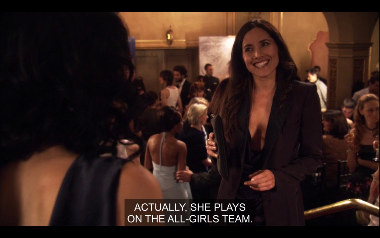 Helena, wearing a high-cleavage black blazer and holding a black clutch purse is smiling in a crowded event hall. She says, "Actually, she plays on the all-girls team."