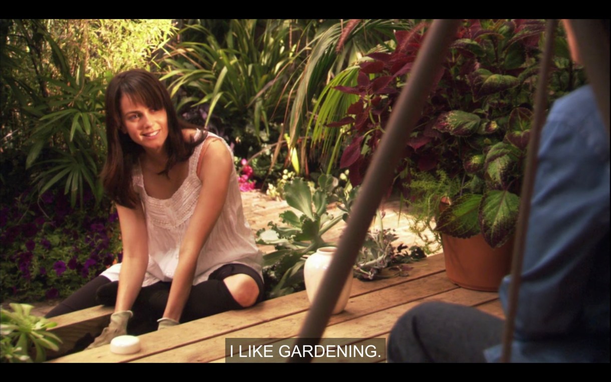 Jenny outside the house gardening in a white frilly tank top. She's looking at Max. We can see Max's back, Jenny says "I like gardening."