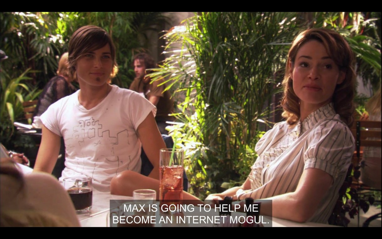 At a table at The Planet with lots of plants behind them. Max wearing a tight white tee sitting next to Alice, who is saying "Max is going to help me become an internet mogul."