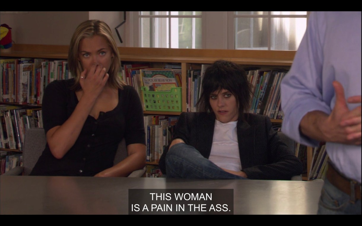 Image Description: Paige and Shane sitting at a table in a school library. Shane's wearing a t-shirt and a blazer and slouching, Paige is sitting up straight with her hand over her mouth. The caption reads, "This woman is a pain in the ass."