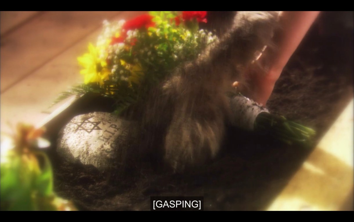 A bouquet of flowers laying next to a small tombstone with the Star of David and "RIP" engraved in it. Subtitles read, "[Gasping]"