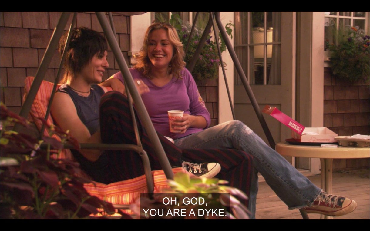 Paige (wearing jeans and a purple top) and Shane (wearing dark pants and a blue tank top) sit next to each other on a porch swing. Shane says, "Oh, God, you are a dyke."