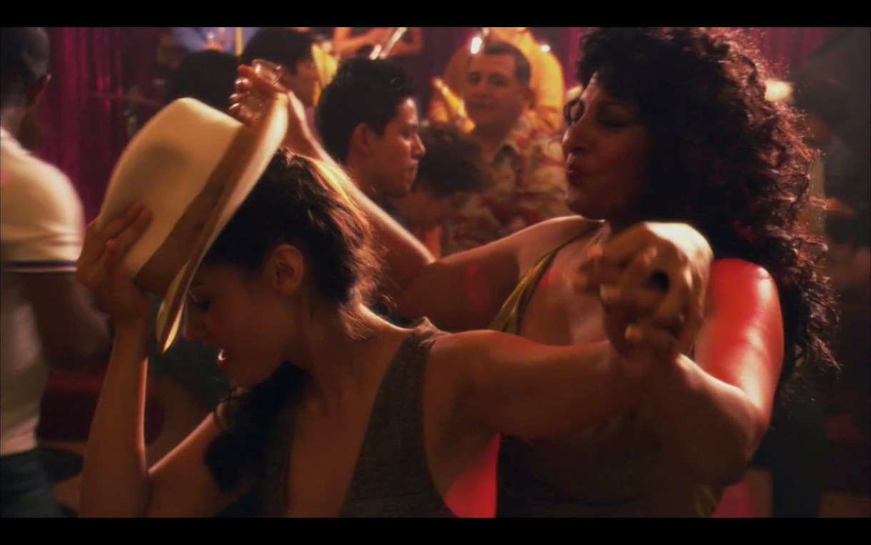 Papi (with a tan fedora on her head) dances in front of Kit, holding hands. There are people around them dancing as well.