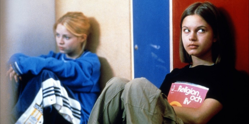 A still from the 15th best lesbian movie of all time Show Me Love. Two girls sit next to each other on the floor with some distance between each other. 