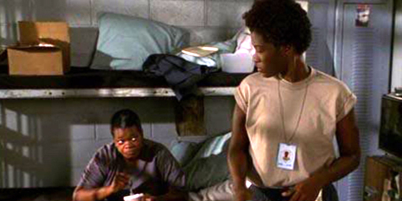 A woman looks down at another woman sitting on a prison bed.