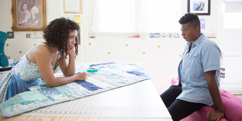 Jasika Nicole and Brittani Nichols stand across from each other looking serious and leaning on furniture.