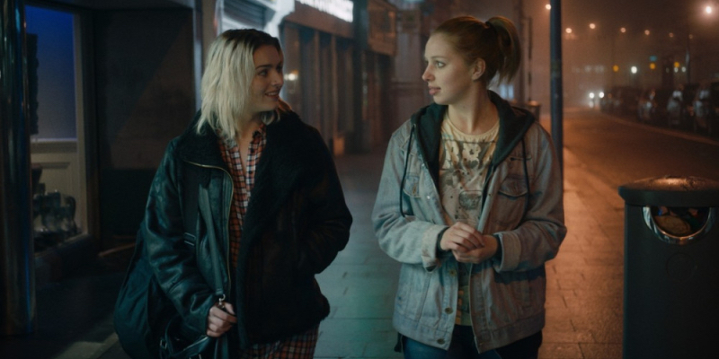 Two young women walk along a street at night.