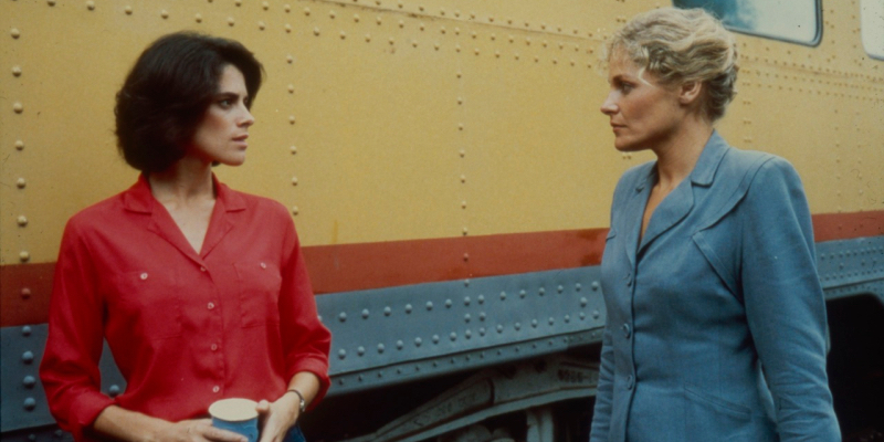 A still from Desert Hearts. Two women look at each other next to a train.