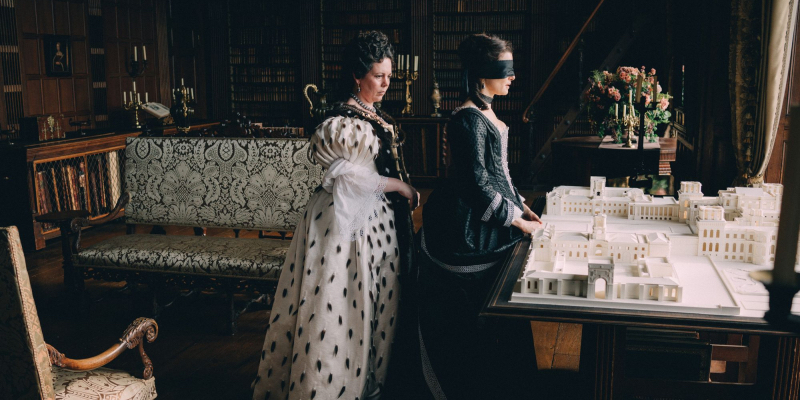 Olivia Colman dressed like a queen stands behind a blind-folded Rachel Weisz.