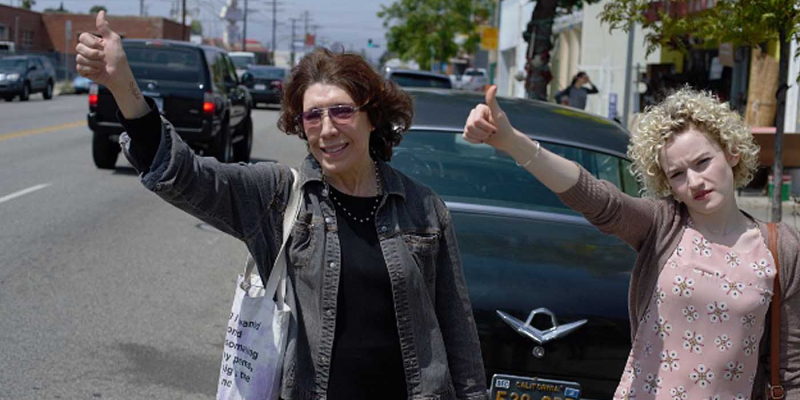 Lily Tomlin and Julia Garner lift their thumbs up on the side of the road.