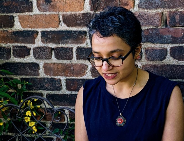 Himani sits on a wrought-iron bench against a brick wall, looking down.