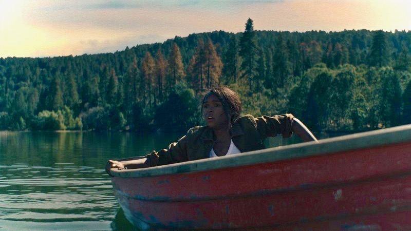 Image: A large lake surrounded by tall, vibrantly green trees. The character played by Janelle Monae is inside a red rowboat. She appears alarmed. She is wearing a white shirt and a green jacket, and clutching both sides of the boat, like she doesn't know where she is or how she got there.