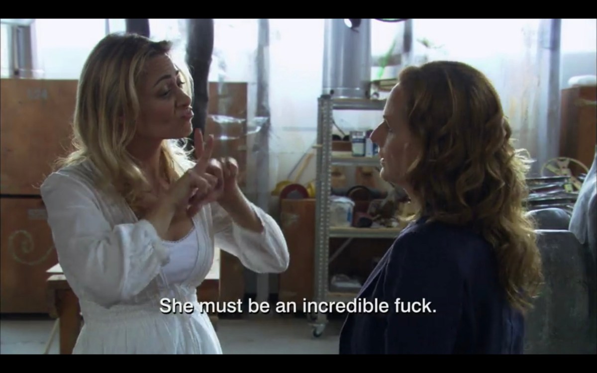 A blonde woman in a white top is signing with Jodi in Jodi's studio. Subtitles read, "She must be an incredible fuck."
