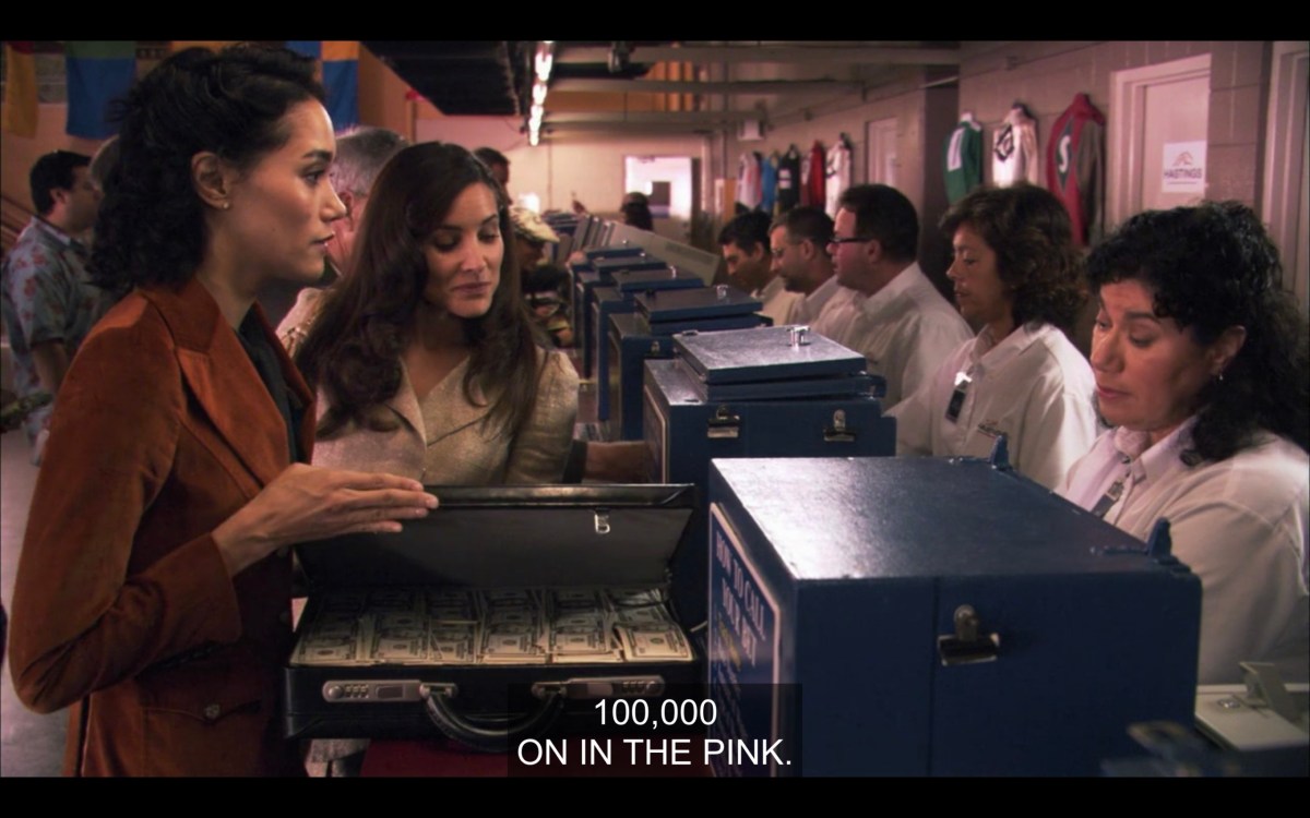 Catherine and Helena at the horse races, about to hand over a briefcase full of cash over to the workers to place a bet. Catherine says, "100,000 on in the pink."