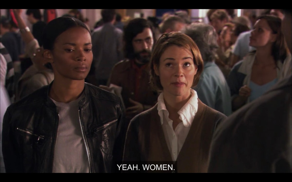 Tasha (wearing a leather jacket) and Alice (wearing a white top and brown cardigan) stand in a crowd at the horse races.  Alice says, "Yeah. Women."