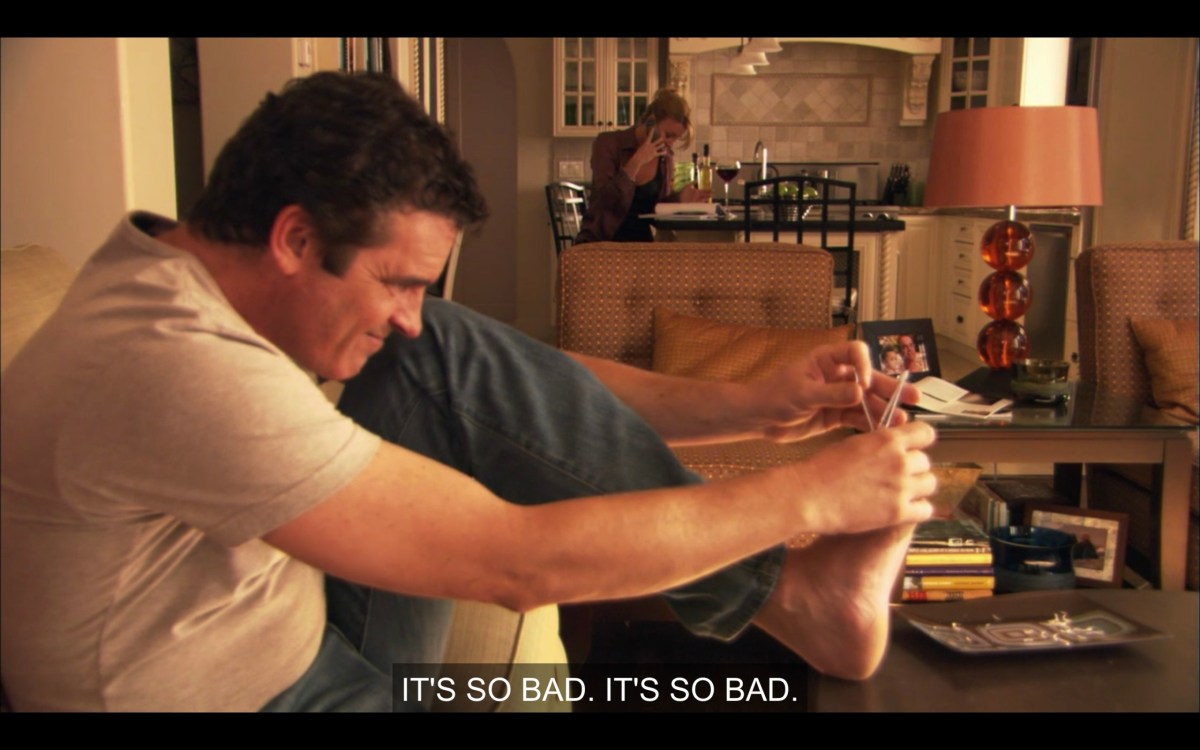 In the foreground, Henry is clipping his toenails on the coffee table. Behind him, Tina is hunched over the counter talking on her cell phone. She says, "It's so bad. It's so bad."