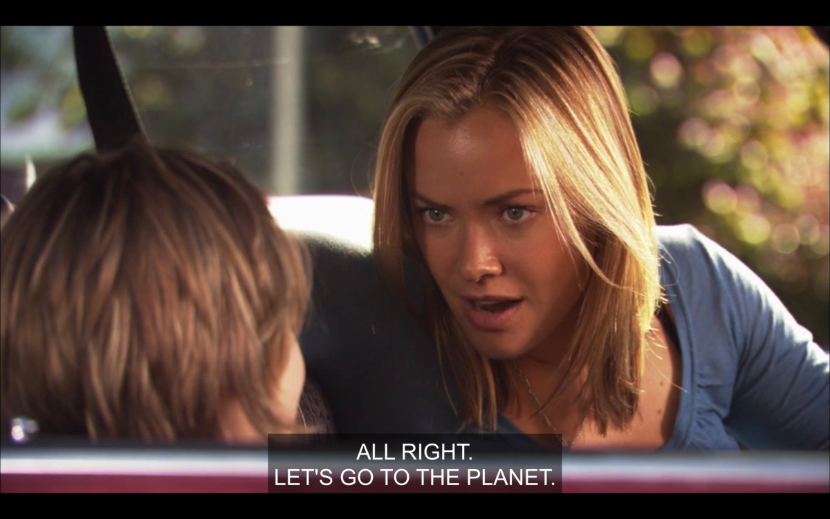 Paige (in a blue shirt) and Jared in the front seat of their car. Paige looks over at Jared and says, "All right. Let's go to The Planet."