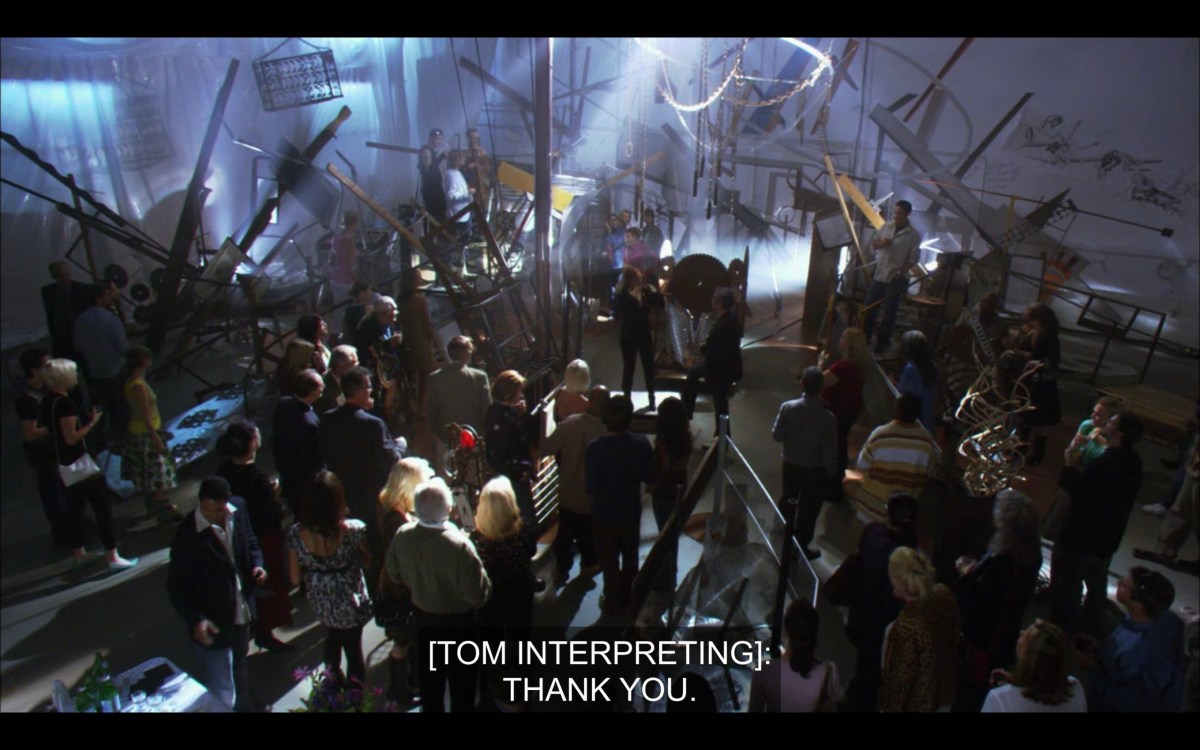 Overhead shot of a crowded room at Jodi's art show. Subtitles read, "[Tom interpreting]: Thank you."