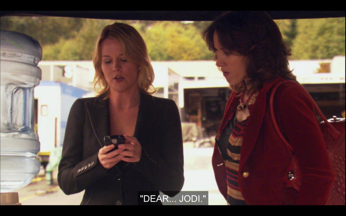 Tina and Bette stand outside next to a water cooler. Tina is looking at her cell phone in her hands, and Bette is looking at it too. Tina says, "Dear... Jodi."
