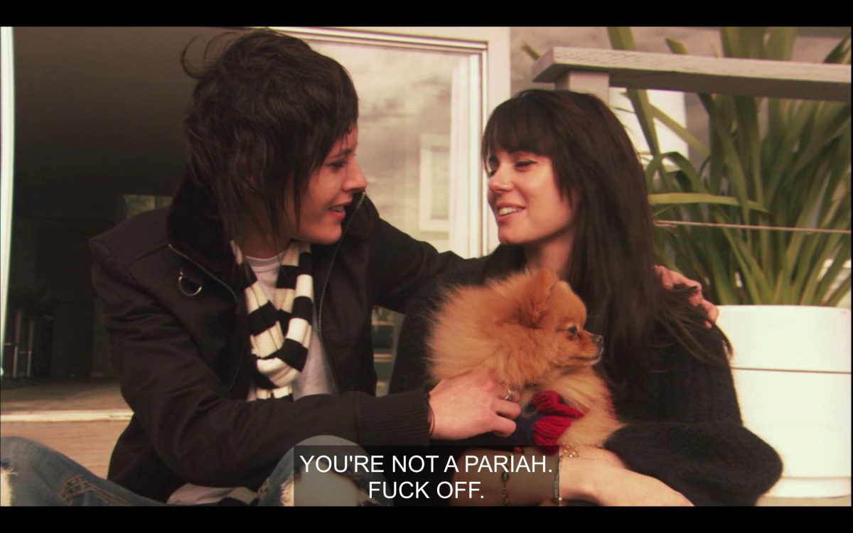 Shane and Jenny (holding a tan Pomeranian dog) sit next to each other outside. Shane has one arm on jenny's shoulder, the other hand on the dog. They are looking and smiling at each other. Shane says, "You're not a pariah, fuck off."