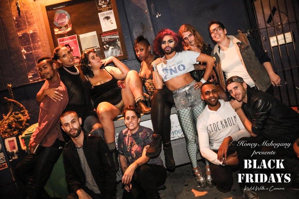 11 queer folks pose for the camera in two rows outside a bar at night; their arms are thrown around each other and they look comfortable with each other. The bottom right corner reads Honey Mahogany Presents Black Fridays. 