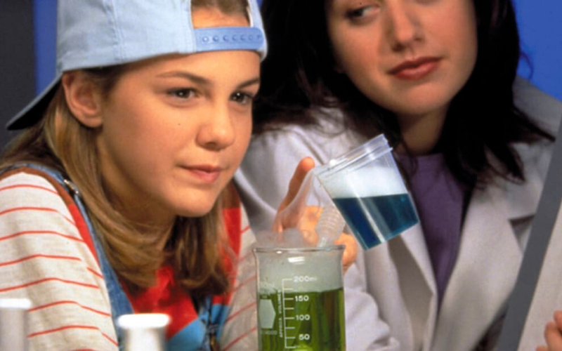 A preteen white girl in a backwards baseball cap looks at a beaker filled with green liquid