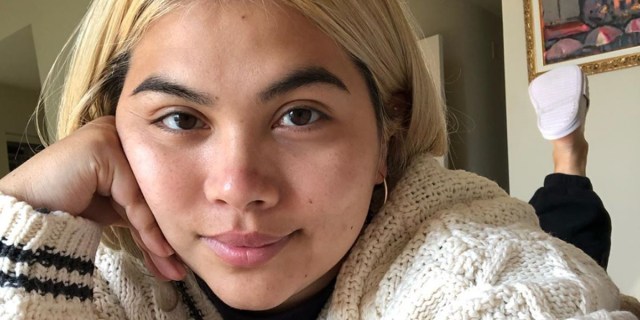 Hayley Kiyoko laying on a bed, looking cozy in a cardigan like the Taylor Swift song.