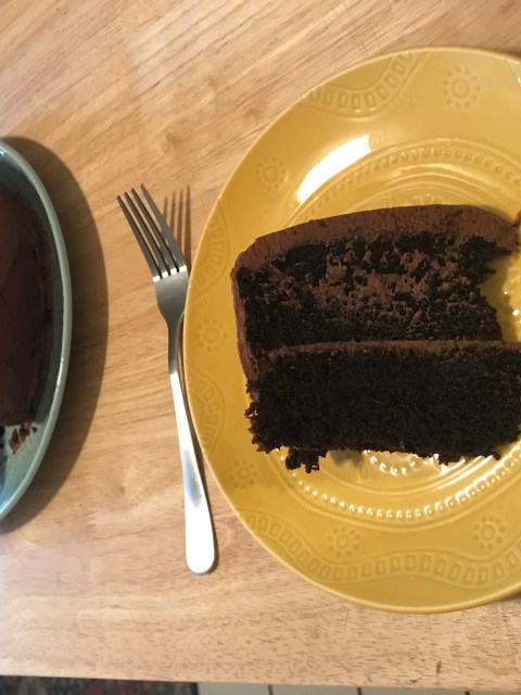 A homemade double chocolate layer cake. In the foreground is a slice of the cake on a yellow ceramic plate with a silver fork. In the background is the full cake (with the slice missing) on a light mint green plate. Both plates sit on a long wooden table.