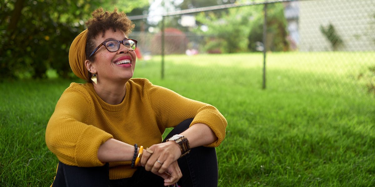 Kirsten Harris-Talley, who is running for public office in Seattle, sits on green grass in a yellow sweater and matching beanie, while she looks up into the sunlight.