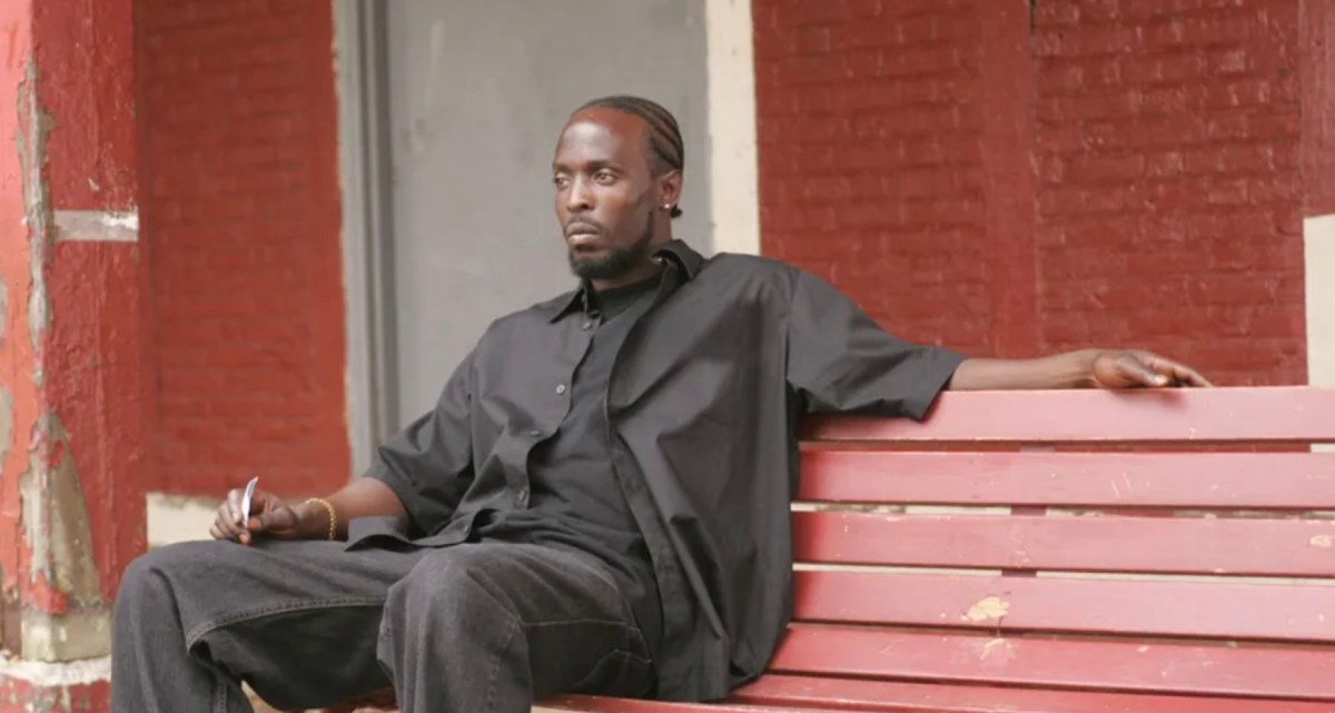 Omar LIttle sits on a red bench in front of a red brick building, wearing all black.