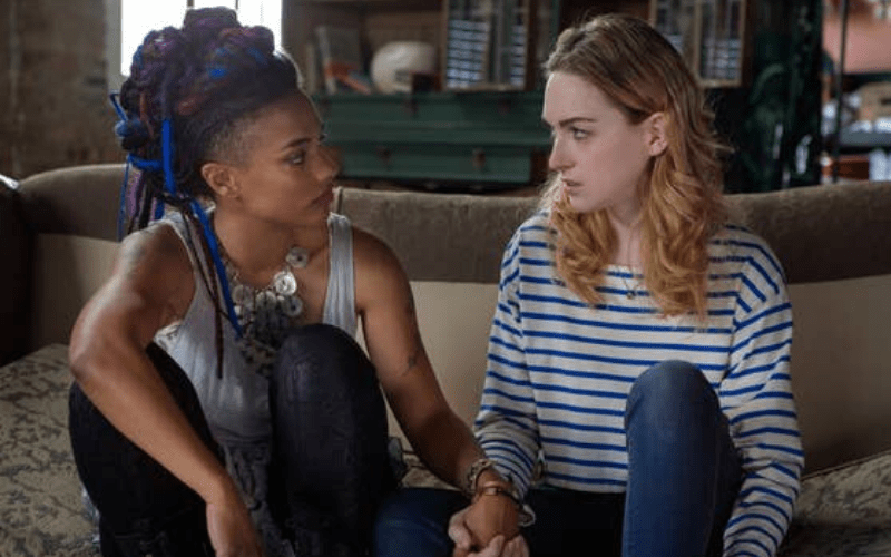 A Black woman with purple and magenta hair holds the hand of blonde, white woman in striped shirt.