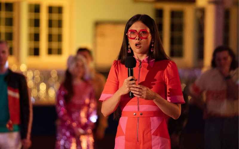 A woman with long brown hair wears a bright pink dress and pink glasses and holds a microphone