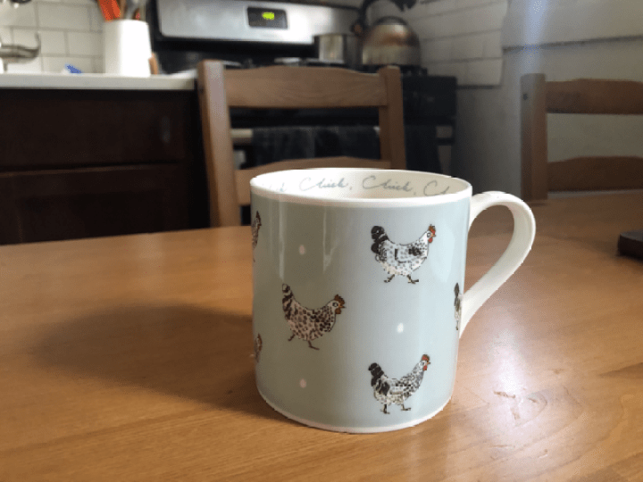 An image of Rachel's mug, sitting on her kitchen table. The table is wood and the mug is a light blue with fancy drawings of chickens all over. The sunlight cascades over the cup and table.