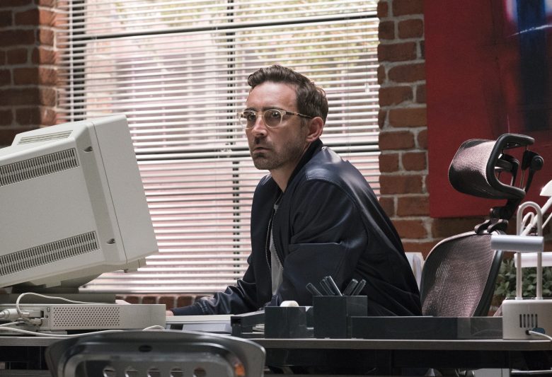 Joe MacMillion sits at a desk in front of a large computer, in the 90s. He's wearing a leather jacket and glasses. The walls are exposed brick, it's the big loft office space.