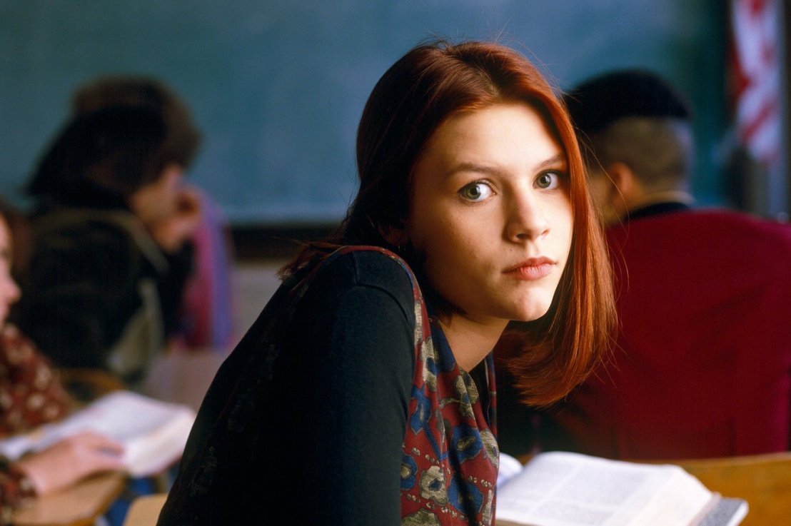 Angela Chase, a teenager with red hair in the mid-90s, stares at the camera in a classroom. It's blurry behind her but you can still recognize ricky and brian's backs.