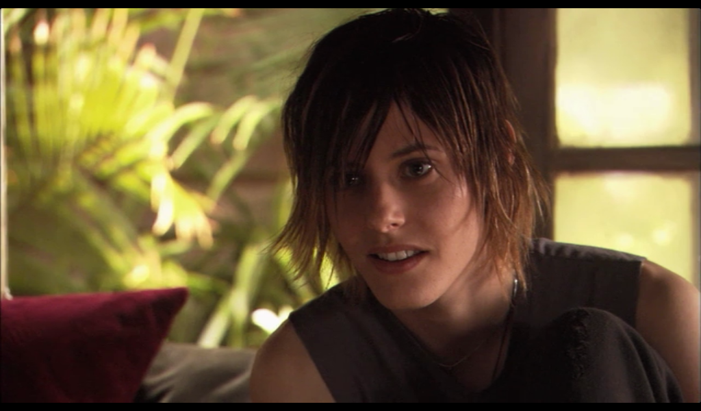 Image: Shane, a lesbian in a muscle tee with a very 2007 haircut, looks at somebody who is out of frame. The L Word.