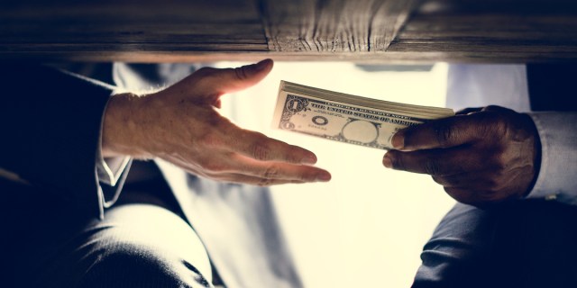 Two hands of people wearing business suits are visible; one is passing the other a stack of bills secretly under a tabletop.