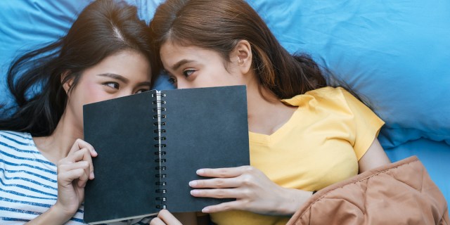 two women with light sexual tension reading together on blue sheets with their faces obscured by the same book