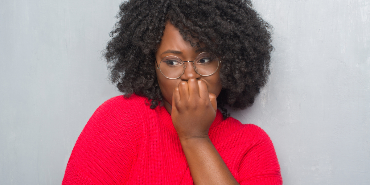 A worried-looking Black woman with natural hair and glasses looking off to the side while she holds her hand to her mouth pensively.