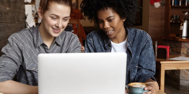 Two women sit in a coffee shop looking at a computer screen. A woman with brown skin and black curly hair holds a coffee cup and smiles while a woman with white skin and reddish brown hair on the left also looks at the computer and smiles. The woman on the left wears a collared shirt, the woman on the right, denim. They give off queer vibes.