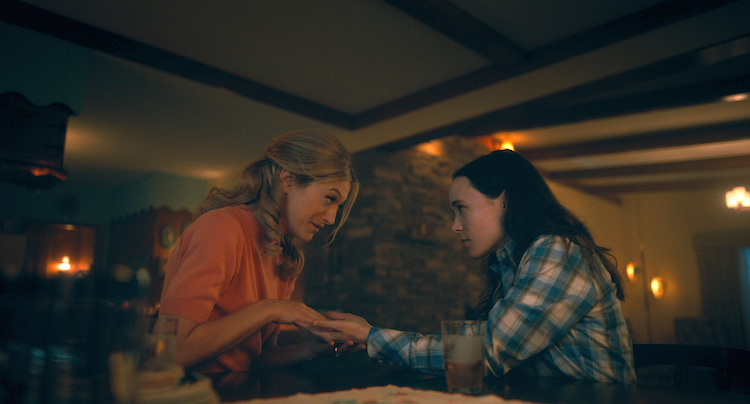Sissy, a blonde woman in an orange shirt, holds the hands of Vanya (Ellen Page), who is wearing a flannel and looking at her intently. They're in a dark but expansive room lit by numerous candles.