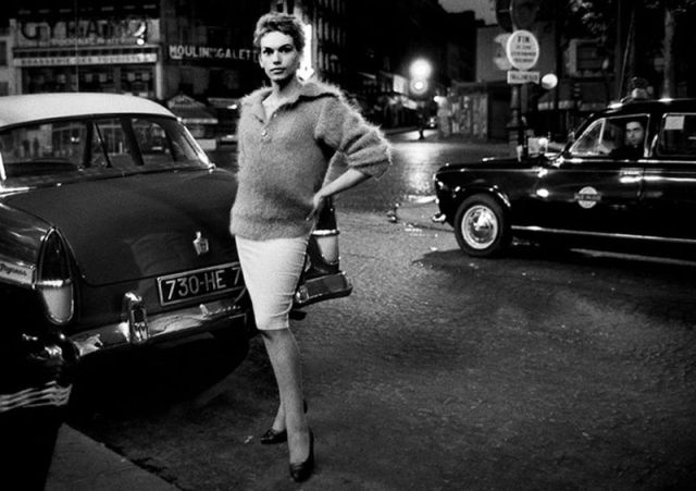 A transgender woman in 1950s/1960s Paris stands in the street at night, her hand on her hip, in a fuzzy sweater and tight pencil skirt. She has her hair pulled back and is wearing a pair of black high heels. It is glamour personified.