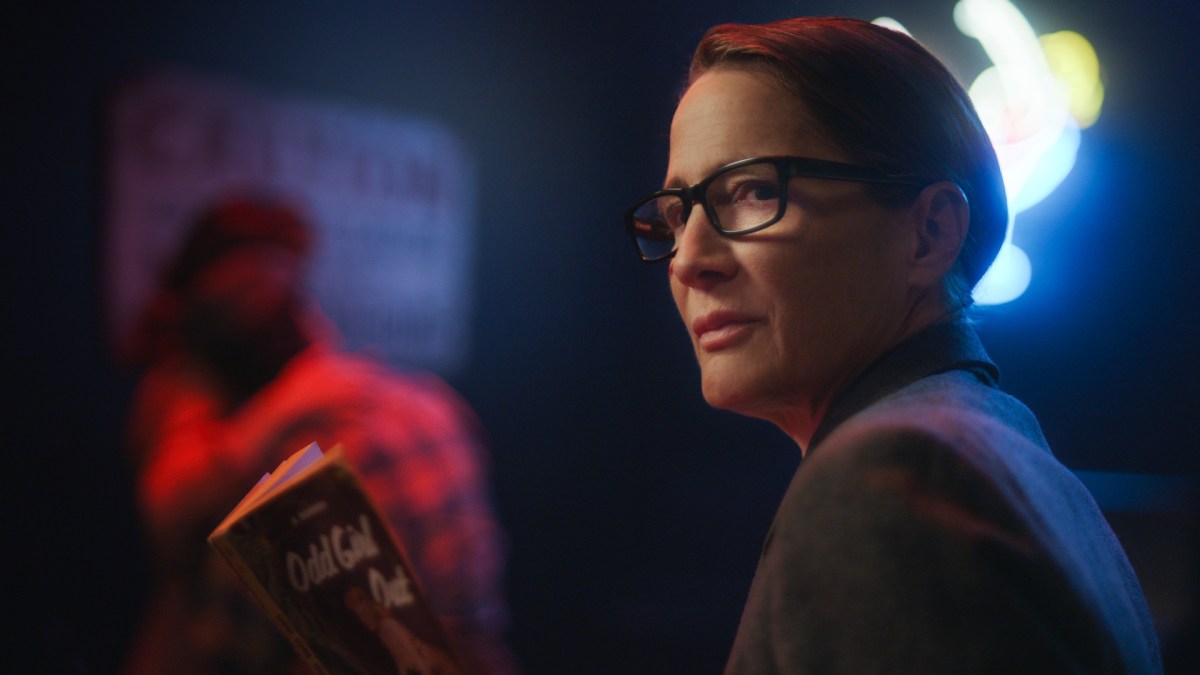 Anne Ramsay as an FBI agent, wearing a ponytail with glasses, reading "Odd Girl Out" in a dark bar