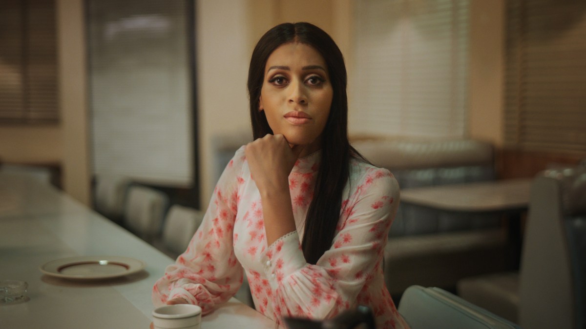 Isis King as Alexis. Sitting a a counter at a cafeteria in a flowered shirt.