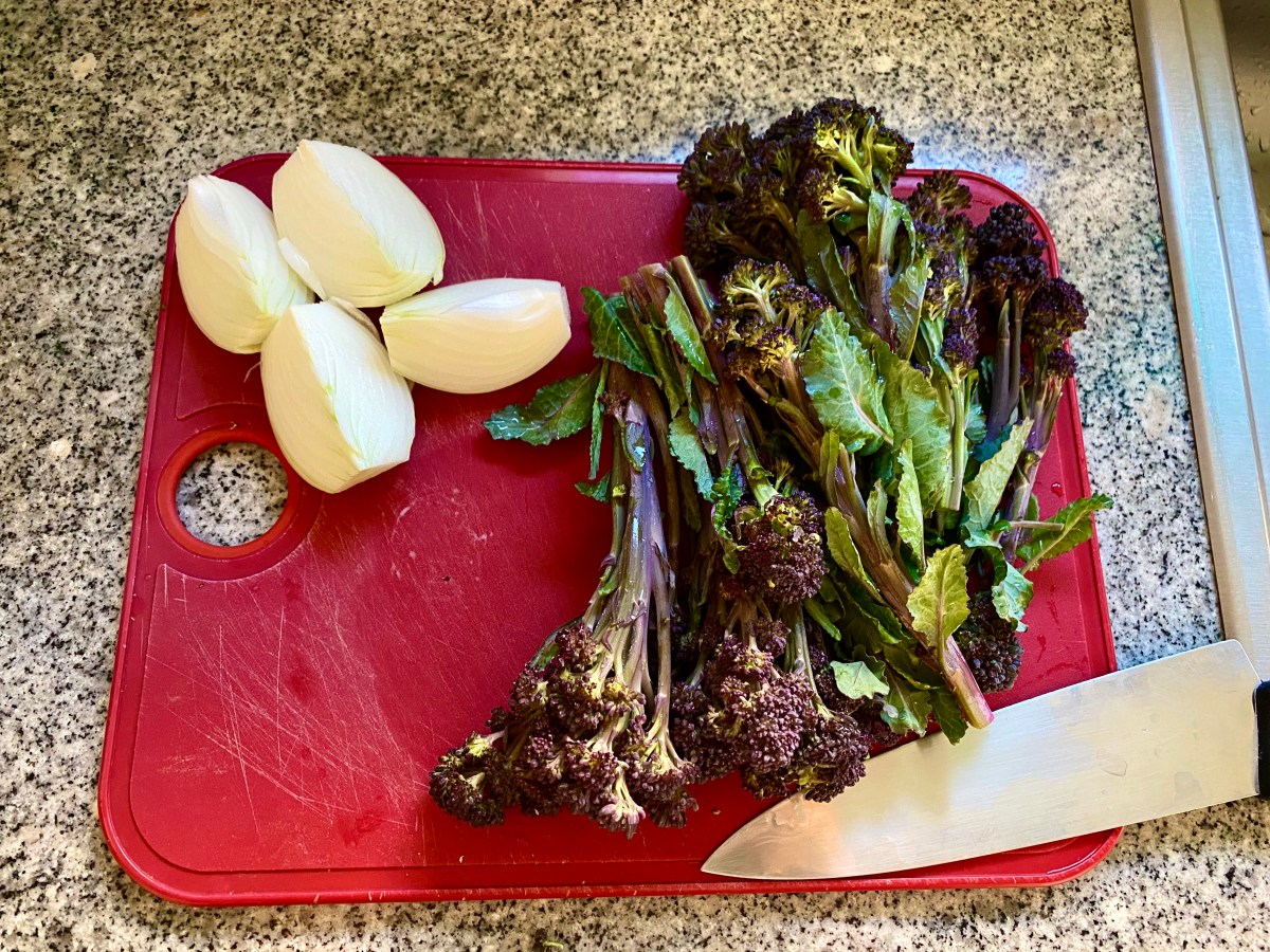 On a red cutting board, a white onion is quartered and two bunches of broccolini with their edges trimmed, are lying next to a knife