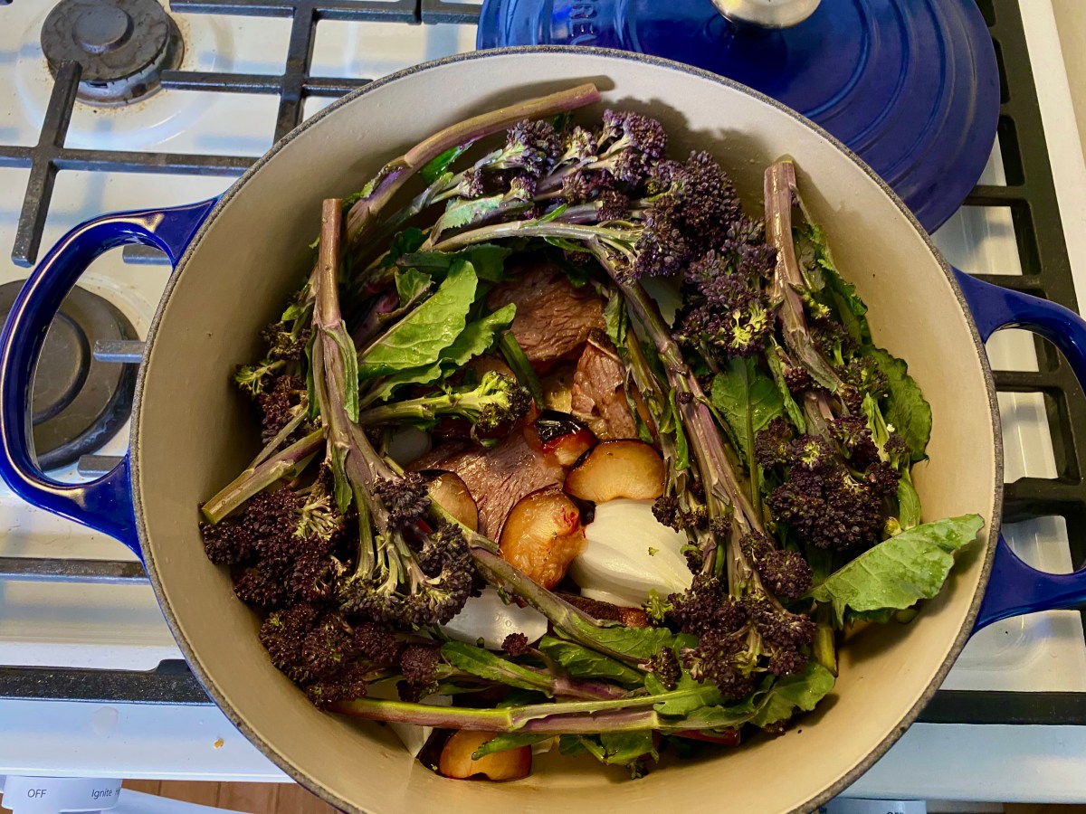 In a dutch oven on top of the stove, beef short ribs are nestled together with plum edges, onion wedges and covered in a wreath of broccolini